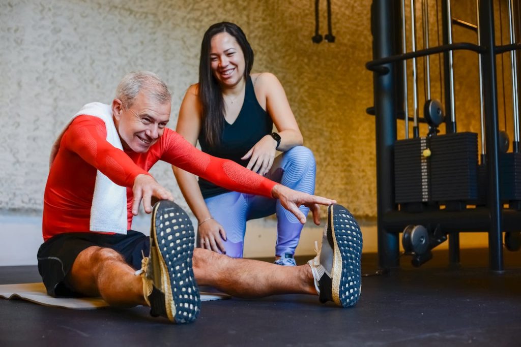 Elderly man in a gym stretching with a fitness coach, promoting a healthy lifestyle.